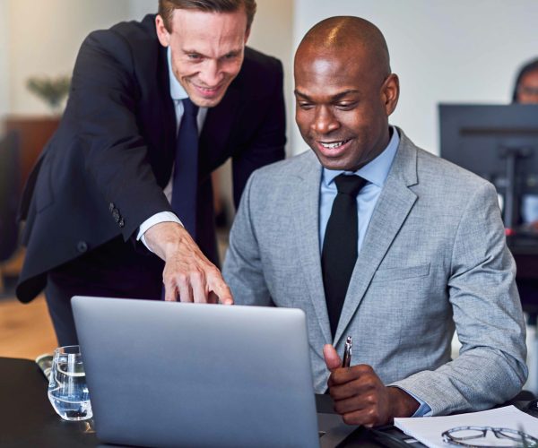 Two smiling diverse businessmen looking at something on a laptop while working together at a desk in a modern office