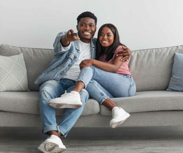 Happy laughing relaxed millennial African American woman and man watching tv in living room interior, holding remote control and hugging. Smiling couple enjoying film and movie evening together